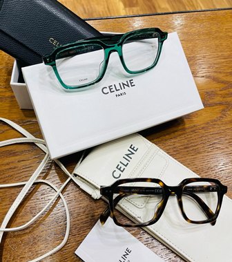 Green-framed eyeglasses resting on a CELINE Paris white box on a wooden table, with another brown frame nearby.