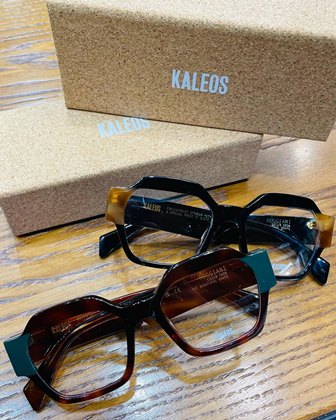 Three Kaleos sunglasses (black, tortoiseshell, and dark brown) on a wooden table with beige boxes labeled KALEOS in the background.