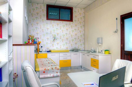 Bright pediatric exam room with yellow-and-white cabinets, a sink, changing table, patterned wallpaper, a small window, and a laptop on a desk.
