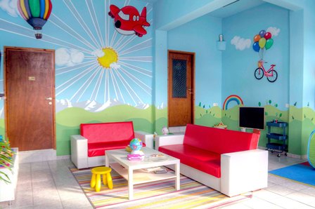 Bright kids' playroom with a blue mural, red-and-white sofas, a white coffee table, and a yellow stool set on a striped rug.