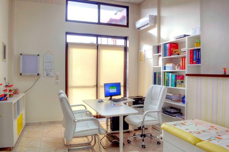 Bright consultation room with a desk, two white chairs, a computer, tall bookshelf, and a large back window.