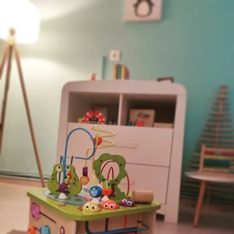 Colorful wooden toy set on a green table in a playroom, with a white dresser and teal wall in the background and a warm lamp