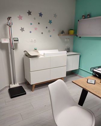 Pediatric exam room with a baby changing station, scale, sink, wall decals of pastel stars, and a white chair beside a wooden table.