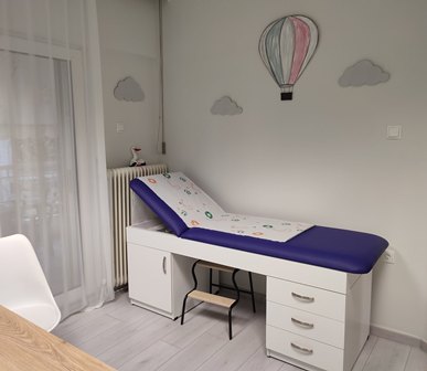 Clinic treatment room with a white examination table, purple cushion, a small step stool, and white storage drawers beneath a radiator; walls have balloon and cloud decals.