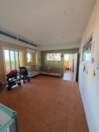 Bright daycare room with strollers and cribs along the left wall, a seating area at the back, and colorful wall decorations.