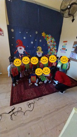 Children pose for a holiday photo in a classroom in front of a Christmas backdrop featuring Santa and a decorated tree.