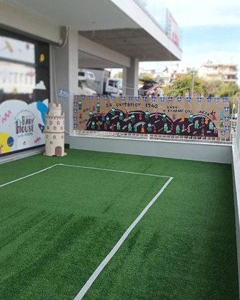 Rooftop play area with artificial grass, white line markings, a beige castle sculpture, and a colorful mural on the wall; city buildings visible beyond.