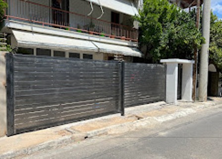 Long dark metal fence and sliding gate along a sidewalk in front of a house, with a white pillar and greenery nearby.