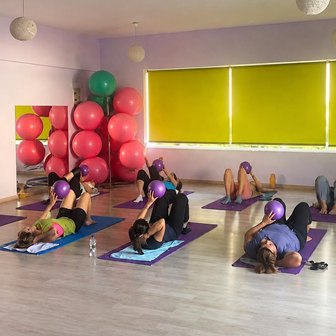 Group of people in a studio doing floor exercises with purple exercise balls; pink balls stacked in the corner and lime-green window blinds in the background.