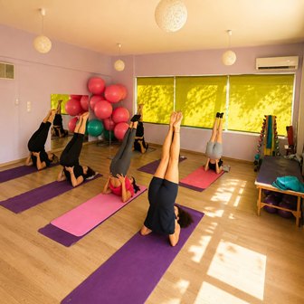 Group of people in a bright yoga studio performing leg-raise inversions on colored mats (purple, pink).