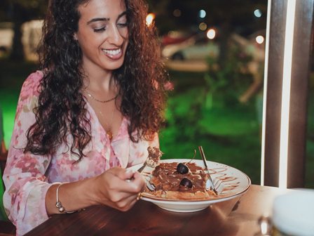 Woman with curly hair and pink blouse smiling as she serves a chocolate pancake topped with berries at an outdoor cafe.