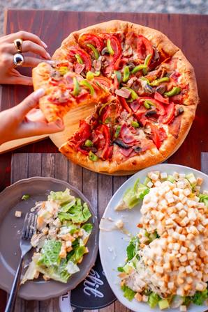 Top-down view of a round pizza with pepperoni, green and red peppers, and melted cheese on a wooden table, with two plated salads nearby.