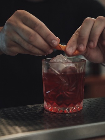 Hands twisting an orange peel over a short glass with a red cocktail and ice, on a bar counter