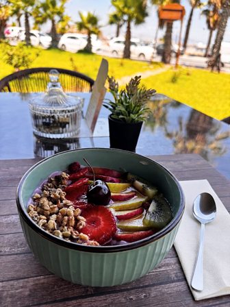 Green bowl of fruit, granola, and yogurt resting on an outdoor wooden table at a sunny cafe; background shows palm trees and parked cars.