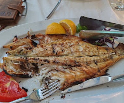 Grilled fish on a white plate with lemon wedges, a tomato slice, greens, and a knife and fork nearby.