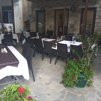 Outdoor restaurant patio with tables set for dining, black chairs, stone flooring, and potted flowering plants nearby.