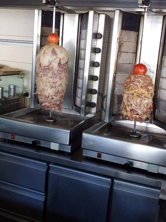 Two vertical rotisserie spits roasting stacked slices of meat, each topped with a tomato, in a kebab shop kitchen