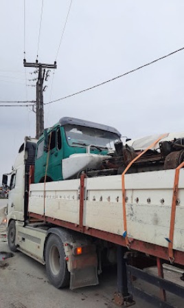 Flatbed tow truck transporting a damaged teal and white truck, secured with orange straps, on a road beside utility poles.