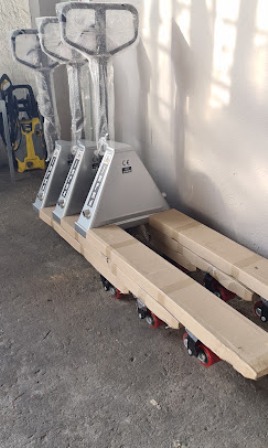 Two pallet jacks leaning against a wall beside long wooden beams on wheeled carts in a warehouse courtyard area.