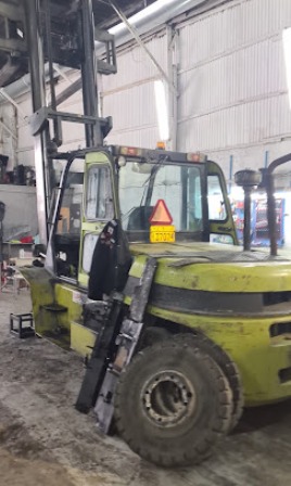 Yellow forklift inside a warehouse, with a ladder leaning against its side and a triangular warning sign on the cab window.