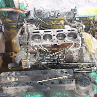 Inline-four car engine block with four piston bores exposed, surrounded by hoses, wires, and a dirty green frame.