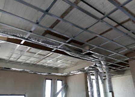 Industrial interior under renovation with exposed metal framing, ceiling grid, and flexible air ducts running overhead.