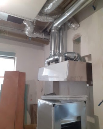 Indoor furnace venting system with large silver ducts connected to a ceiling-mounted hood, in a room under construction.