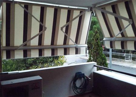 Balcony with striped retractable awnings extended, hose reel on the wall, and a view of a street with greenery beyond.
