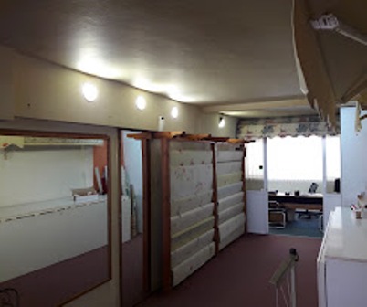 Interior view of a narrow office hallway with frosted glass partitions and wooden dividers, leading to a back room with desks visible in the distance, lit by ceiling lights.