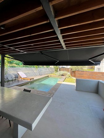 Under a dark shaded patio, a rectangular backyard pool with lounge chairs and a wooden privacy fence.
