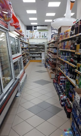 A supermarket aisle with refrigerated units on the left and shelves full of bottles and boxes on the right, bright tiled floor and ceiling lights.