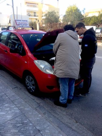 Two men inspect the engine under the open hood of a red hatchback parked on a city street.