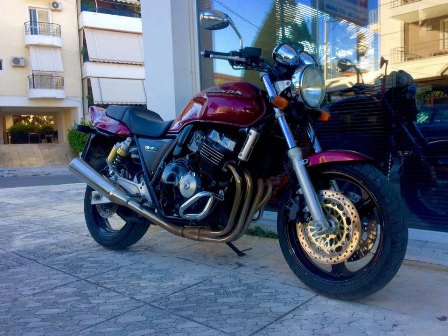 Side view of a maroon motorcycle parked on a sidewalk in front of a glass storefront, showing chrome exhaust and disc brakes.