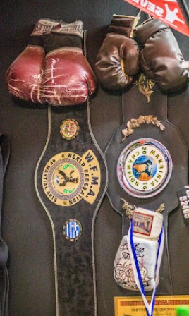 Two boxing/mma championship belts hanging on a wall, with brown gloves resting above them on a dark display surface.