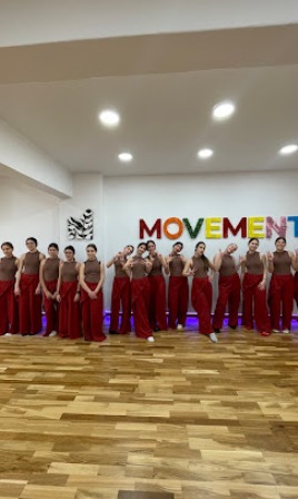 Group of people in red pants standing in a line in a dance studio with a wooden floor and colorful MOVEMENT letters on the wall behind them.