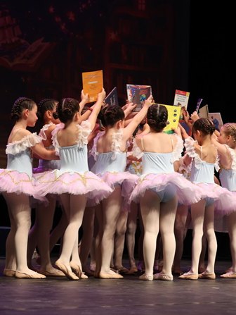 Group of young ballet dancers in light blue leotards and pink tutus form a circle on stage, holding colorful books above their heads.