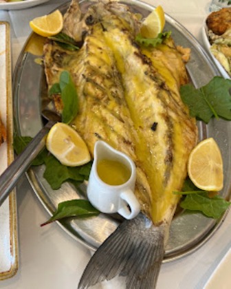 Whole grilled fish on a platter with lemon wedges, greens, and a small sauce pitcher.