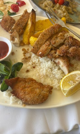 Plate of breaded fried fish, fried chicken, potato wedges, and white rice, with a lemon wedge, greens, and a small cup of red sauce on the side.