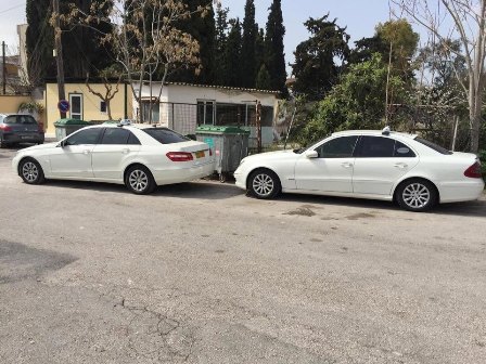 Two white sedans parked on a street beside a green dumpster and a beige building, facing opposite directions.