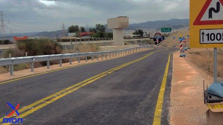 Two-lane road under construction with yellow center lines, metal guardrails, and construction signs; distant workers and hills in the background.