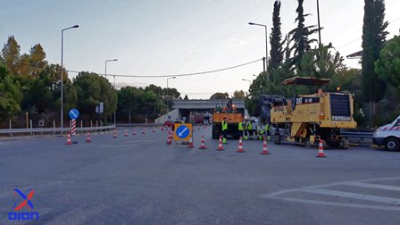 Roadwork scene with orange cones and barriers blocking lanes; workers in high-visibility vests and a large yellow construction machine ready to repair the road on a sunny day.