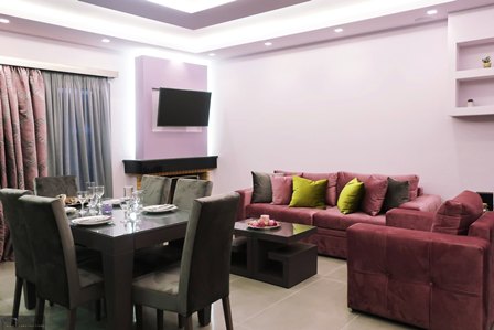 Modern living room with a pink sectional sofa, dark dining table for six, and a wall-mounted TV. Pale purple walls and patterned curtains add color.