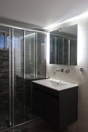 Modern bathroom with a glass shower enclosure, dark vanity, white sink, and large wall mirror beneath bright overhead lighting