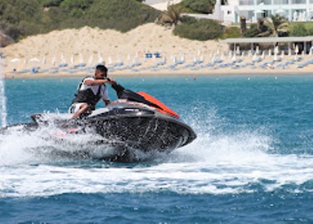 Person riding a black and red jet ski on blue water with a sandy beach and lounge chairs in the background.