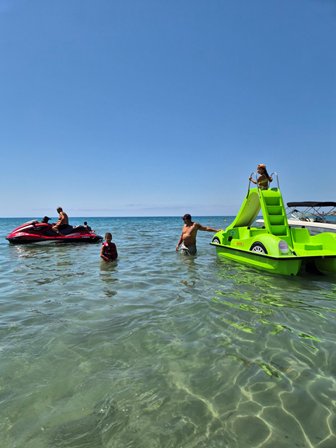 People swimming and standing in clear water next to a bright green boat and a red jet ski on a sunny day by the shore equilibrium of sea and sky.