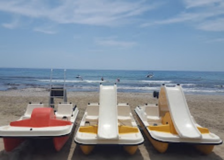 Three pedal boats lined up on a sandy beach with the calm blue ocean and sky in the background, and swimmers visible in the water.