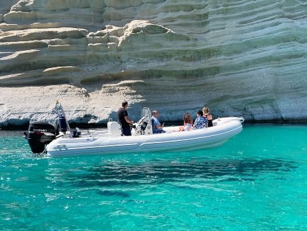 White inflatable motorboat with several people aboard near a rocky cliff and bright turquoise water.