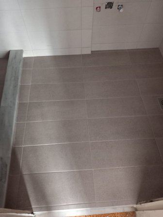 Gray bathroom floor with large rectangular tiles; a step/ledge along the left and a floor drain in the bottom-right corner, suggesting under-construction area.