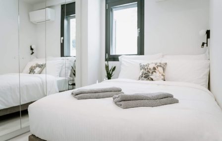 Bright minimalist bedroom with a white bed, two folded towels, decorative pillows, and a mirrored wardrobe reflecting the room.
