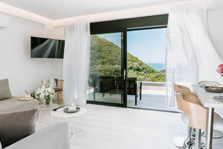 Bright living room with white decor and a glass sliding door opening to a balcony overlooking green hills and the sea.